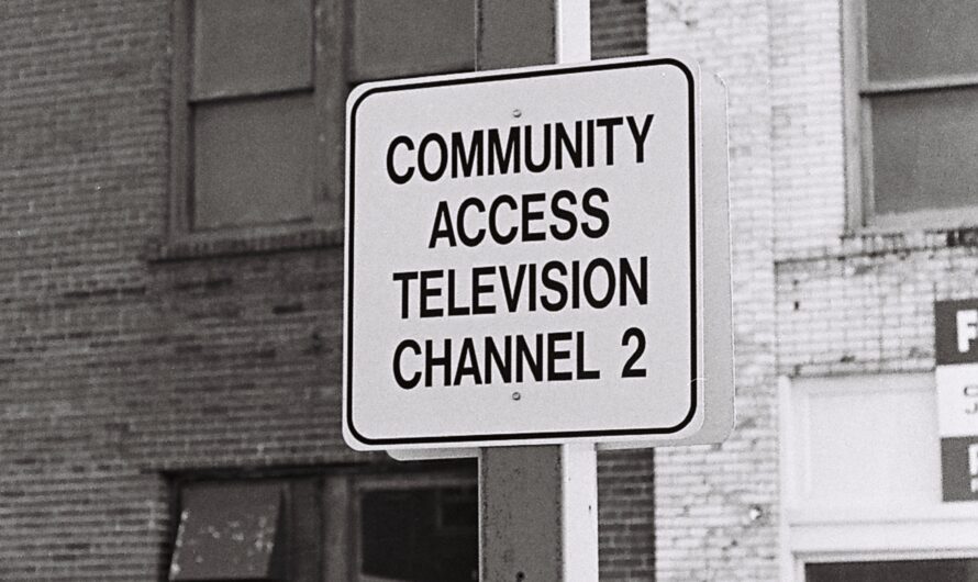 Against the background of two conjoined, many-storied brick buildings, a white sign reading “Community Access Television Channel 2” in all-caps, block, black lettering