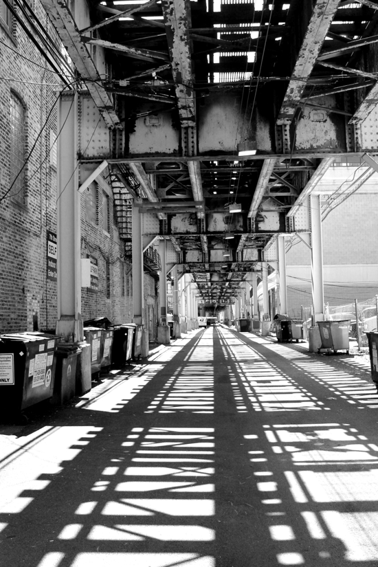 The black and white photo features a view looking down a street under the elevated train. The ground shows the shadow of the track’s pattern. The street is lined with steel supports and dumpsters. 