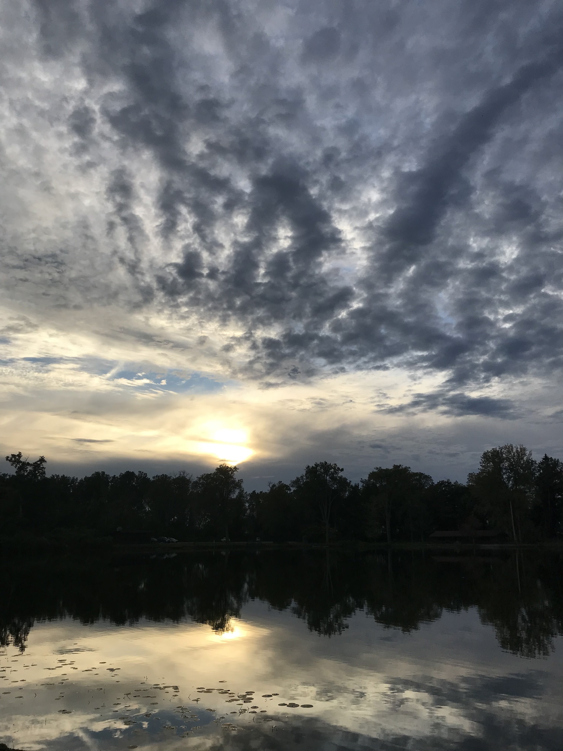 Pictured is a sunset over a lake with a dark tree-line in the distance, reflected in the still water, and scattered clouds in the sky.