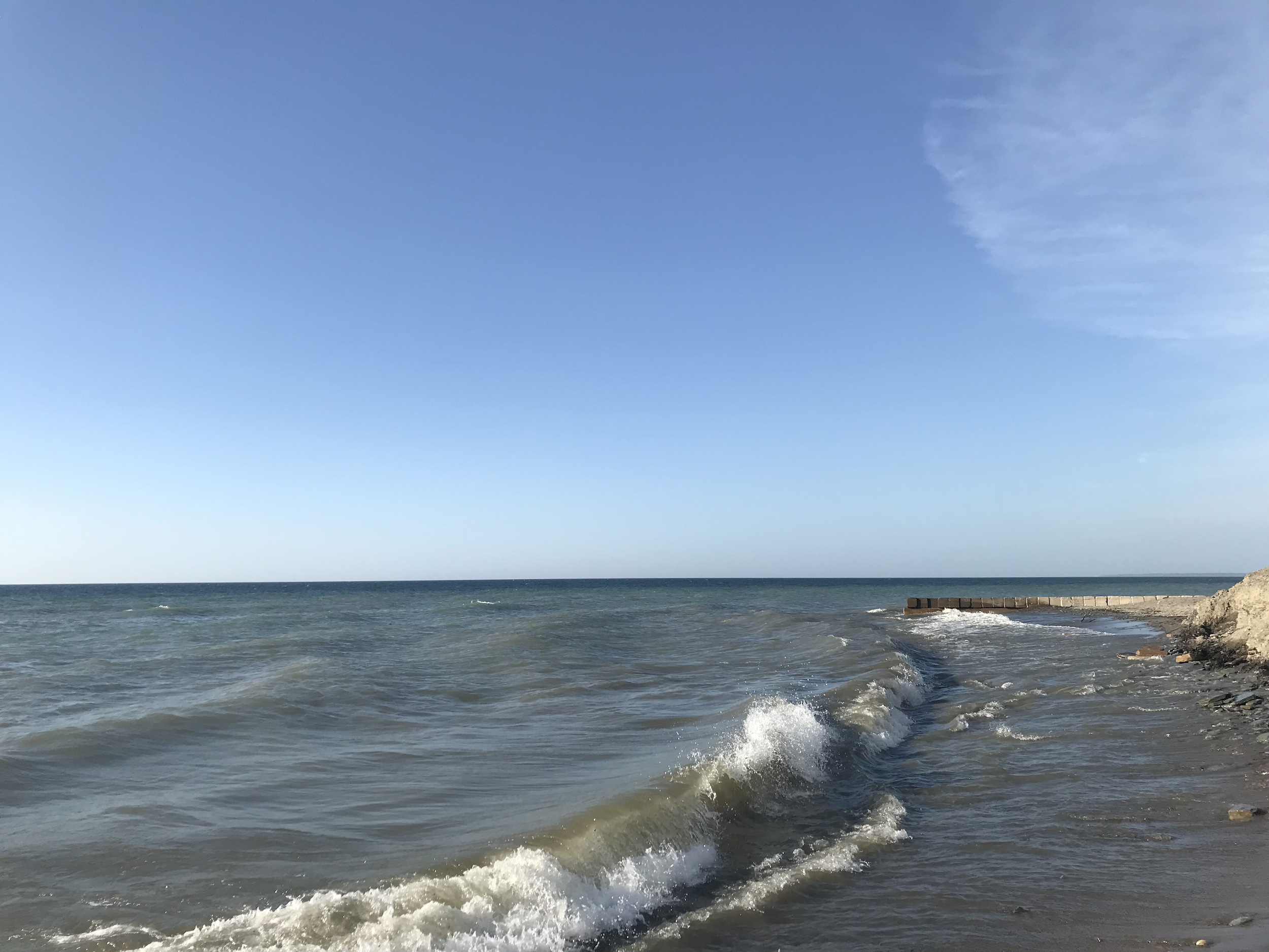Pictured is the Shoreline of Lake Erie with small waves rolling onto the beach and a clear blue sky overhead.
